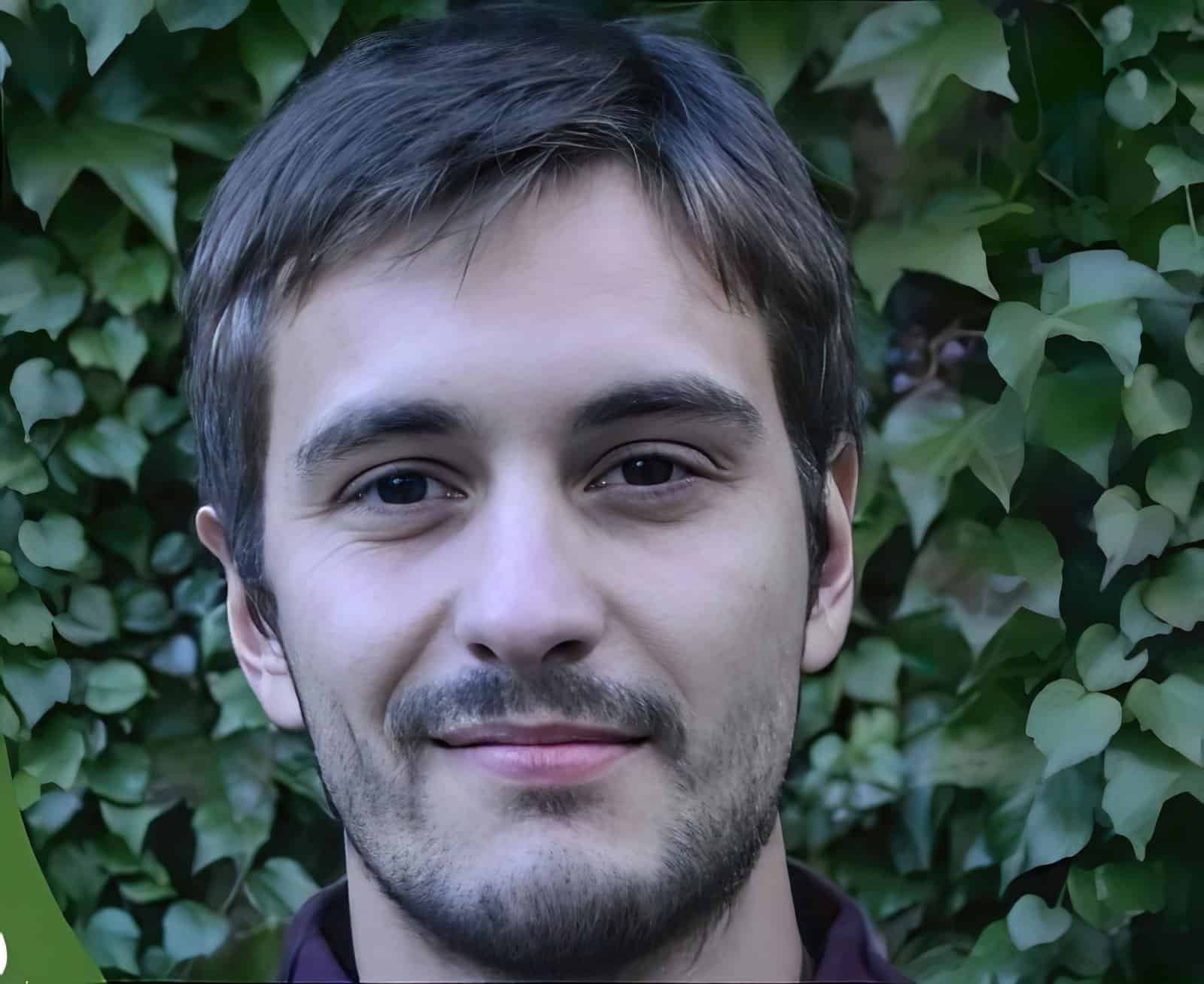 Portrait of a smiling man with short brown hair against a leafy green background.