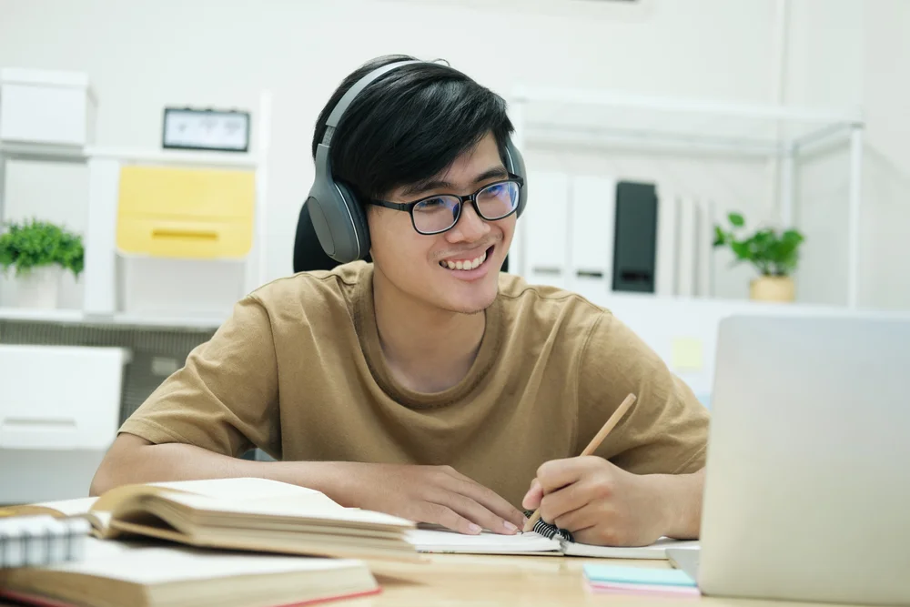 Smiling Asian male student wearing headphones, studying online with laptop and notebook.